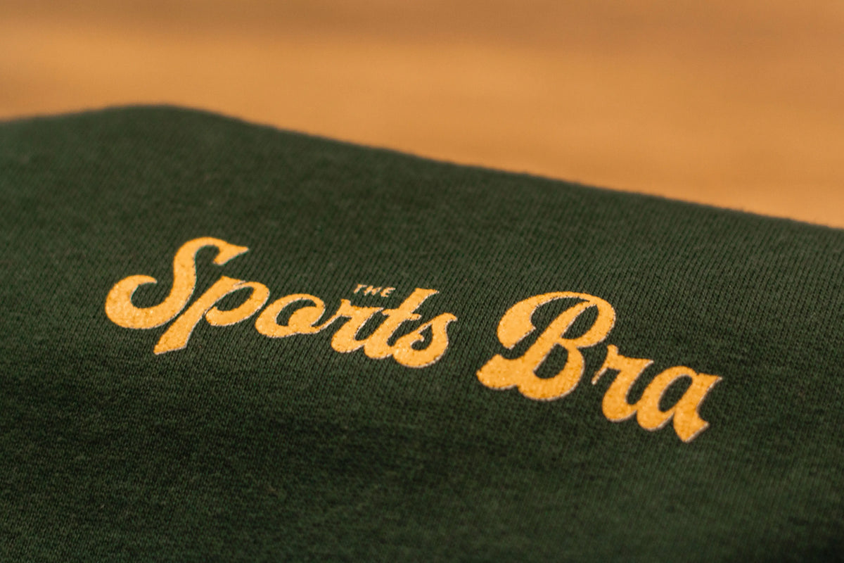 Close-up and detail view of the upper left front design The Sports Bra® signature style font in a warm, golden color. Sweatshirt looks soft, is evergreen color. There is a nice contrast.