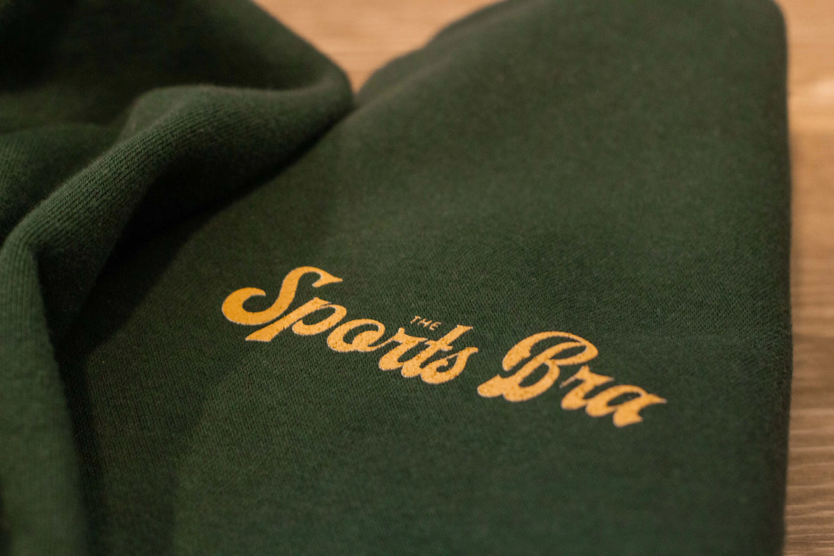 View of the upper left front design The Sports Bra® signature style font in a warm, golden color. Sweatshirt looks soft, is evergreen color. There is a nice contrast. 