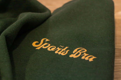 View of the upper left front design The Sports Bra® signature style font in a warm, golden color. Sweatshirt looks soft, is evergreen color. There is a nice contrast. 